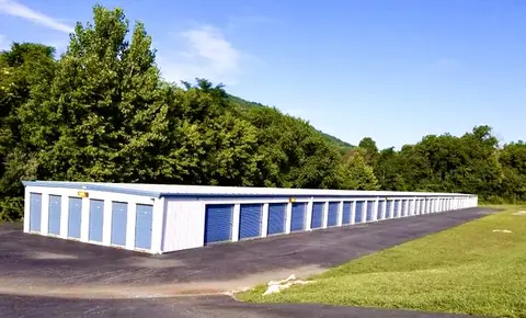 Long white self storage building with blue roll-up doors on a paved drive, backed by trees and Shenandoah Valley hills at American Mini Storage in Staunton, VA
